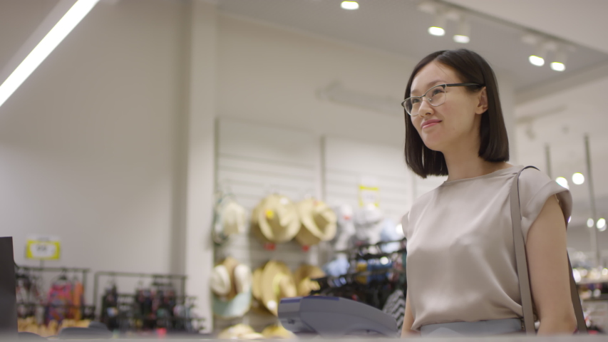 Waist-up shot of smiling Asian woman scanning her palm on payment terminal at checkout counter of clothing store, taking shopping bags from sales assistant, thanking, saying goodbye and leaving Royalty-Free Stock Footage #1039895603