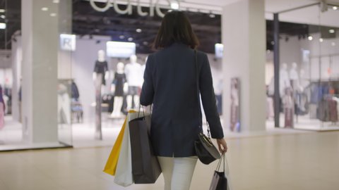 Tracking rear shot of slim young woman in white jeans and blazer with plenty of shopping bags walking through mall and entering brightly lit clothing chain store with mannequins on display
