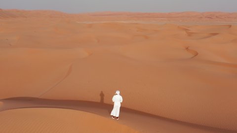 Rising drone shot of man in traditional arab clothing (thoub) stands on a dune in a desert. Crane style shot with camera tilt down. Warm sunset colors with long shadows