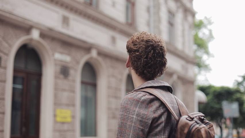 Young Millennial in Glasses Wearing Checked Coat and Jeans in Earphones with Bag Walking, Looking around at Old City Street. Student, Tourist Concept. Traveling.