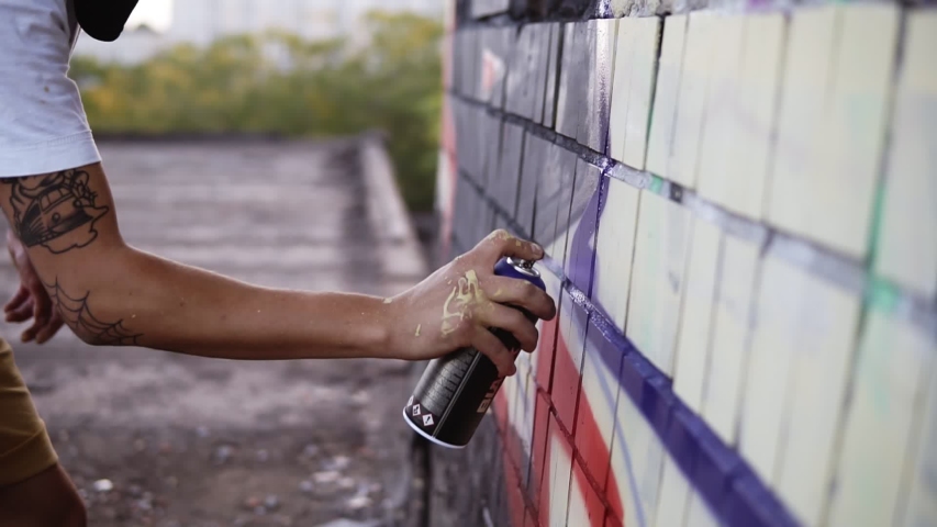 Close-up of artists hand dirty in paint applying spray drawing a blue coloured lines on a street building wall. Action. Stained fingers of artist hold spray can with colored paint on concrete wall