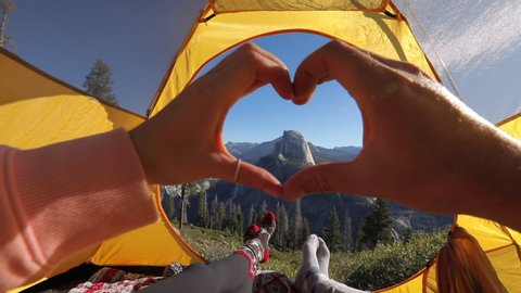 A couple has a day out in Yosemite Valley, admiring the beauty of Sierra Nevada mountains and the Half Dome rock from the tourist tent. They make a hand heart against the backdrop of the landscape.