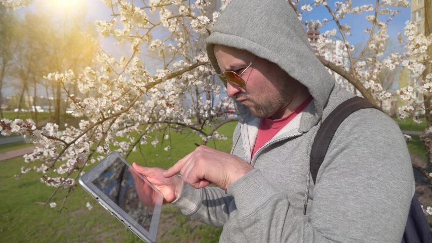 Adult man plays the game on tablet pc. Angry man with tablet PC stands near apricot tree in city park