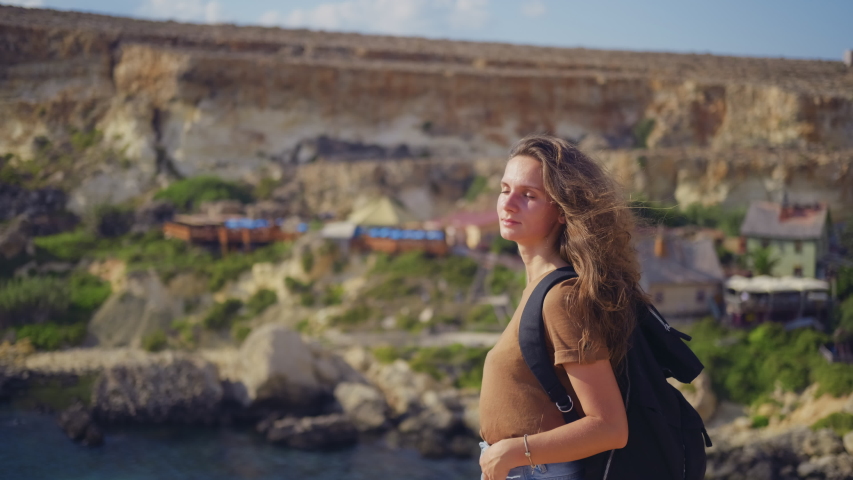 Young woman tourist with a black backpack walks on the rocks with views of the sea and the village.
