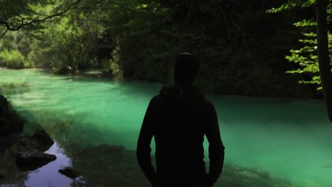 Back view man standing in Beautiful background of Turquoise colorful waterfall cascade river lake in Spain in summer. Blue transparent water, and green forest. Pebbles under the water. 4K UHD.