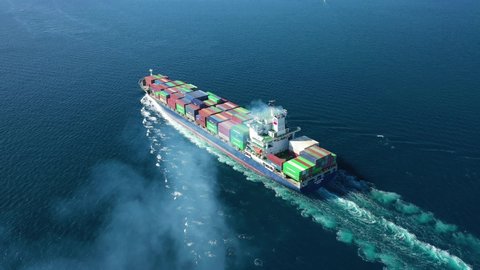 Aerial drone video of Container cargo Ship carrying load in truck-size colourful containers cruising deep blue open ocean sea 