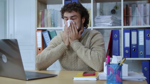 LONDON, UNITED KINGDOM, 20 OCTOBER, 2019:Tired caucasian office worker sneezing from allergy blowing nose using napkins working on laptop computer in the company. Hardworking businessperson suffers