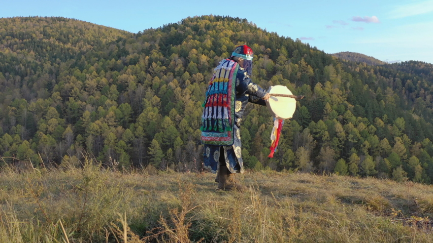 Energetic dance of an old shaman with a tambourine on the background of autumn mountain landscape.