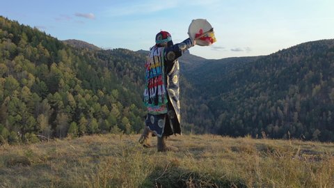 Energetic dance of an old shaman with a tambourine on the background of autumn mountain landscape.