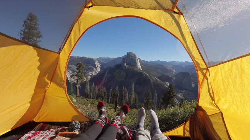 Two pairs of legs in the opening of the tent, against the backdrop of the mountain landscape of Sierra Nevada and the cliff of Half Dome.The sun illuminates the tent.Outside landscape looks inspiring.