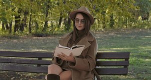 Smart elegant Caucasian woman in sunglasses and brown hat sitting on bench and turning pages of the book. Beautiful European girl reading poetry in the autumn park. Cinema 4k footage ProRes HQ.