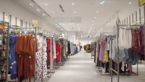 Dollying shot of camera moving through aisle in fashion store and passing by racks of carefully arranged brightly colored clothing prepared for opening