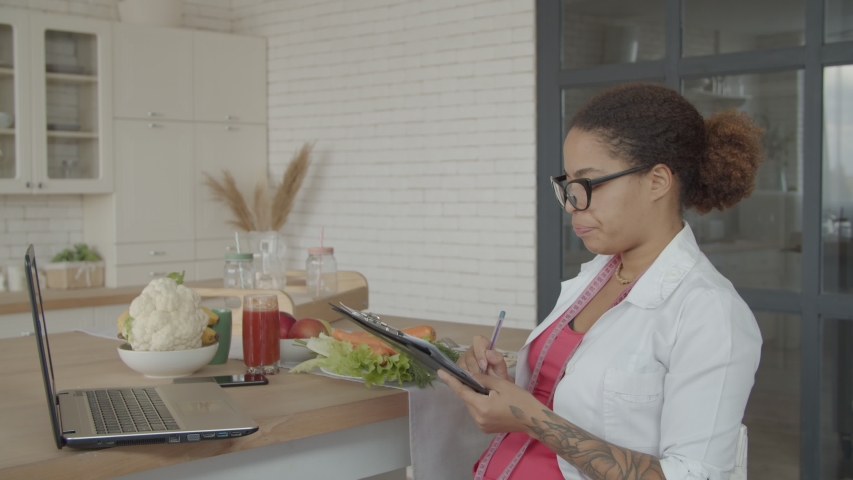 Positive african american female nutritionist in eyeglasses and uniform having a phone call and scheduling appointment in clipboard. Cheerful woman dietitian consulting patient using smartphone.