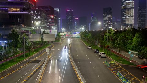 Timelapse wide Nanshan district highway with cars with busy traffic between green trees and illuminated Shenzhen city buildings at dark night