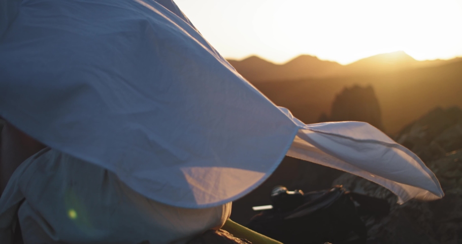 Young woman at sunset covering warm blanket blowing in wind on mountain top. Back of delighted girl tourist warming up observing beautiful evening sky and nature scenery.