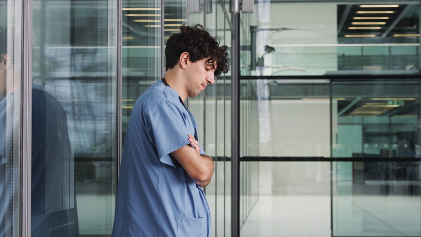 Tired Young Male Doctor Wearing Scrubs Sitting Against Wall In Modern Hospital Building Royalty-Free Stock Footage #1039963208