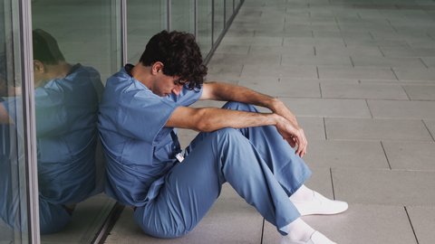 Tired Young Male Doctor Wearing Scrubs Sitting Against Wall In Modern Hospital Building
