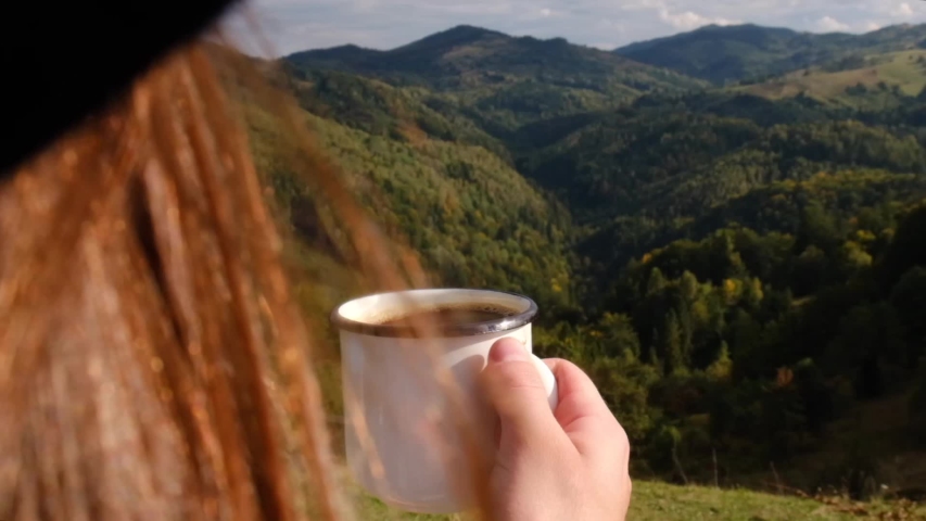 View from over shoulder on female traveler holds lifts a cup of coffee demonstrating overlook amazing of mountains landscape, beautiful and inspiring camp location for outdoor vibes living lifestyle