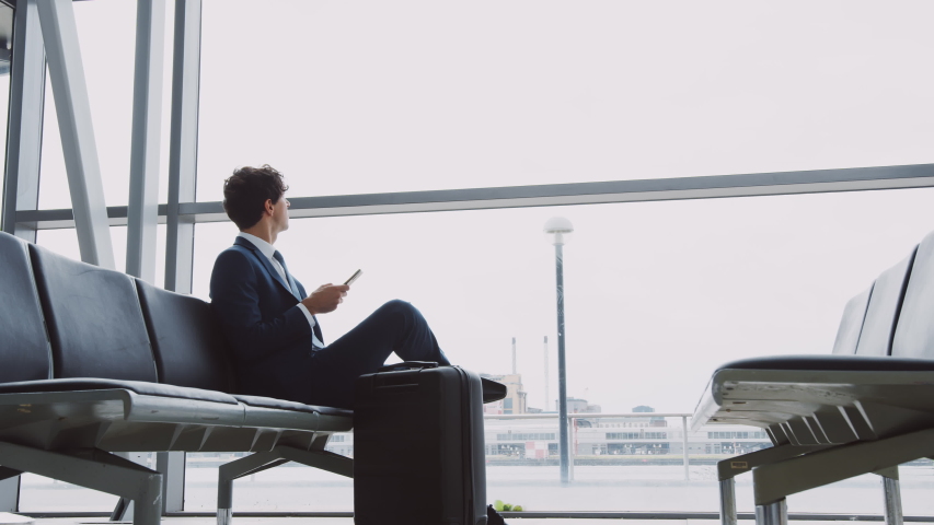 Businessman Sits In Airport Departure Lounge Using Mobile Phone With Plane Taking Off In Background Royalty-Free Stock Footage #1039964774