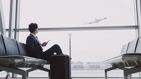 Businessman Sits In Airport Departure Lounge Using Mobile Phone With Plane Taking Off In Background