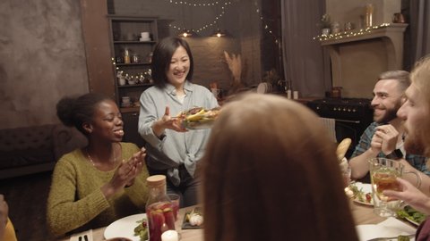 Handheld shot of happy Asian woman smiling and putting dish of roasted steak with vegetables on dinner table, then clanking glasses with group of cheerful friends and celebrating