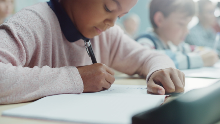 In Elementary School Classroom Brilliant Black Girl Writes in Exercise Notebook, Taking Test. Junior Classroom with Diverse Group of Bright Children Working Diligently and Learning. Portrait Royalty-Free Stock Footage #1039971821