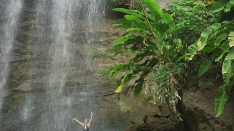 Tilt down to distant happy woman standing on rocks under waterfall / Carmel Falls, Grenada