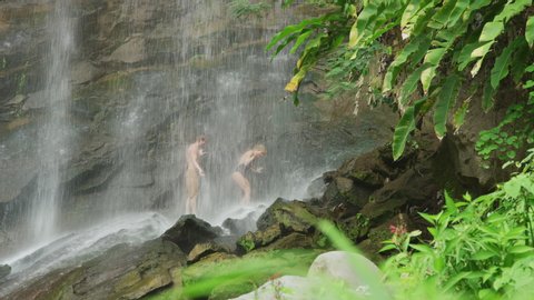 Tilt down to couple walking on rocks under waterfall / Carmel Falls, Grenada