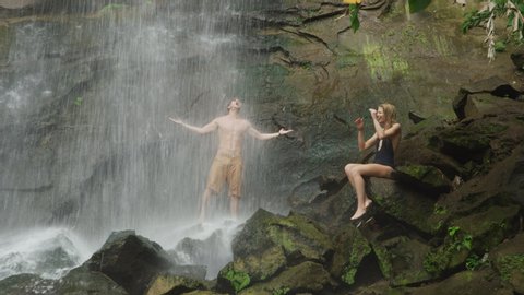 Woman sitting on rock watching water splashing on man standing under waterfall / Carmel Falls, Grenada
