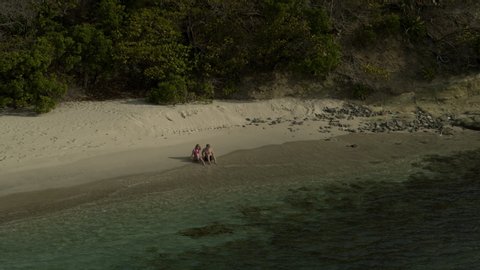 Aerial zoom in to couple sitting on ocean beach near waves / Anse La Roche bay, Carriacou, Grenada