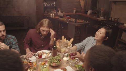 Handheld tracking shot of group of happy people smiling and clanking glasses at dinner party in cozy home