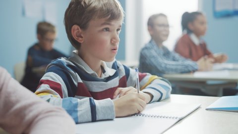 In Elementary School Class: Portrait of Brilliant Caucasian Boy Writes in Exercise Notebook, Taking Test and Writing Exam. Diverse Group of Bright Children Working Diligently and Learning