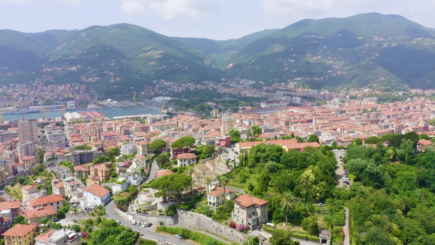 La Spezia, Italy. A sweeping aerial view reveals the city nestled between the Apennine Mountains and the Gulf of Poets, with terracotta rooftops cascading down the hillside toward the historic port. The camera glides over the Poggio hill, where the medieval Castello San Giorgio once anchored the Genoese defense of the bay, offering a panoramic perspective of the dense urban grid, the naval harbor, and the deep natural inlet that has drawn mariners since Roman times. Lush Mediterranean vegetation frames the residential quarters, while warm summer light bathes the Ligurian coastline in rich, saturated tones. The scene captures the layered character of a working maritime city that has served as both a strategic naval stronghold and a gateway to the Italian Riviera.