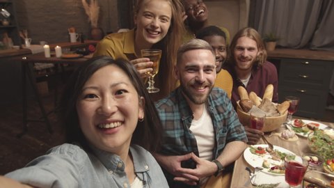 POV shot of happy Asian woman laughing and taking selfie with friends at dinner party in her cozy home