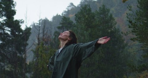 Lovely young girl with wet curly hair in green raincoat looking up enjoying rainy day in nature, Hiker Woman Raises her Hands Feeling Freedom and Unity with Nature in Autumn Forest