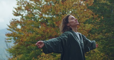 Happy young woman in green raincoat looking up with raised hands enjoying autumn rainy day, feeling of freedom 