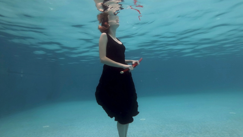 Surreal girl slowly sinks under the water to the bottom of the pool in a black dress with a red samurai sword in his hand. She pulls out her sword, shows it, and plays with it. Portrait. Slow motion.