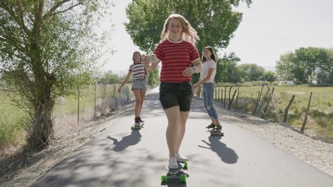 Tracking shot of girls riding skateboards on park path posing for cell phone selfies / Saratoga Springs, Utah, United States