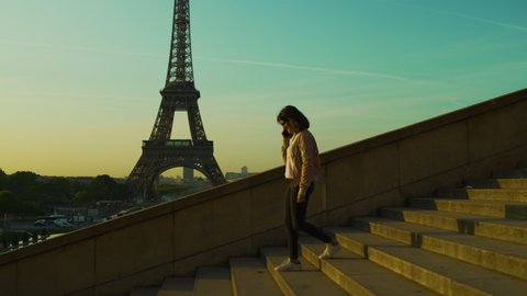 Attractive Girl having phone conversation and laughing going down stairs with smartphone at Eiffel tower during sunset orange blue sky in Paris in the summer. Trocadero. Steadicam side track 4K UHD. Stockvideó