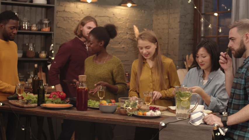 Handheld medium shot of group of friends drinking wine and chatting while cooking dinner in kitchen. Cheerful young man with beard and long hair smiling and snacking on grapes Royalty-Free Stock Footage #1039988648