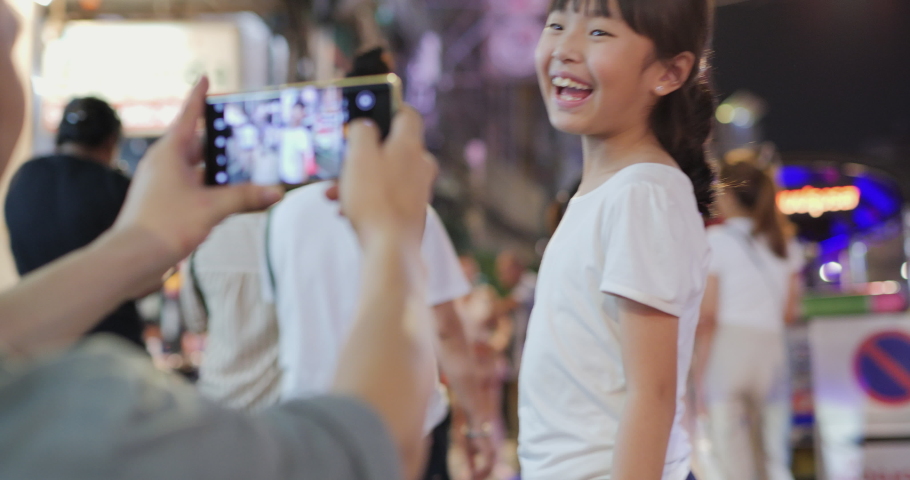 Asian little girl posing for portrait photo at city. Father using smartphone camera to take little girl photo.