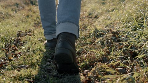Close-up man legs/feet steep terrain on field in evening rays of sun, steadicam shot. Male hiker traveler in jeans and boots making steps, hiking, autumn season trekking, stepping on fallen leaves