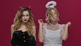 Emotional happy positive women playing with a hair demon and angel in carnival costumes isolated over red wall background