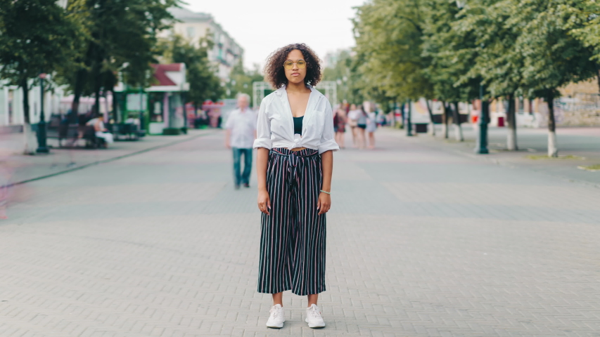Time lapse portrait of attractive African American girl standing outdoors on pedestrian street and looking at camera with serious face. People and lifestyle concept.