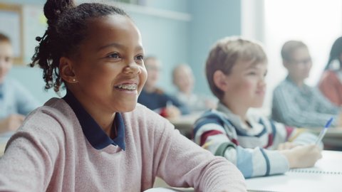 In Elementary School Class: Portrait of a Brilliant Black Girl with Braces Writes in Exercise Notebook, Smiles. Junior Classroom with Diverse Group of Bright Children Working Diligently and Learning 