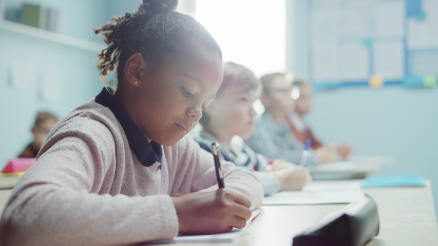 In Elementary School Class: Portrait of a Brilliant and Cute Black Girl with Braces Writes in Exercise Notebook, Smiles. Junior Classroom with Diverse Group of Bright Children Working Diligently | Shutterstock HD Video #1040038283