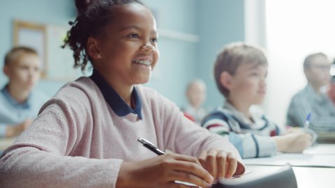 In Elementary School Class: Portrait of a Brilliant and Cute Black Girl with Braces Writes in Exercise Notebook, Smiles. Junior Classroom with Diverse Group of Bright Children Working Diligently