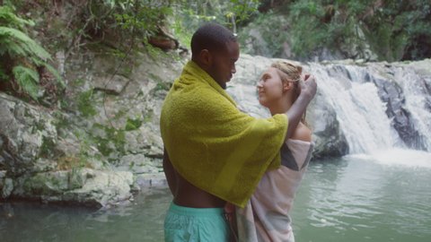 Young beautiful couple wrapped in towels holding each other at a secluded river near waterfall. Medium close shot on 4k RED camera.