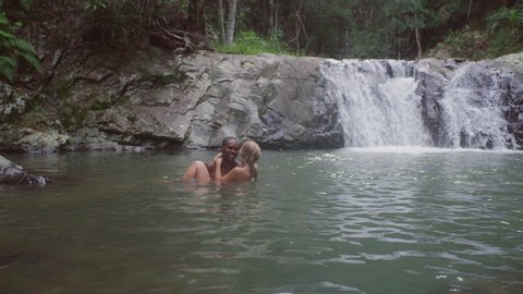 Young beautiful couple going swimming holding each other in secluded river near waterfall. Wide shot on 4k RED camera.