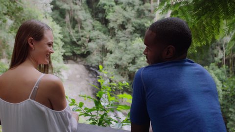 Two millennial friends leaning on railing smiling in front of small waterfall in an Australian rainforest by safety railing during daytime. Medium shot with zoom on 4k RED camera.